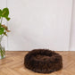 A Round Natural Sheepskin Pet Bed in brown, eco-friendly and hypoallergenic, lays on a wooden floor from Mellow Pet Store.